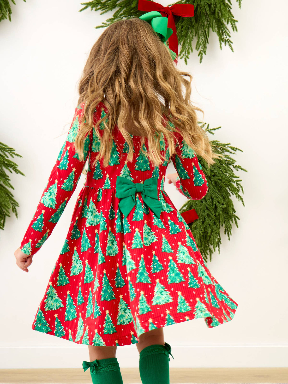 A young girl viewed from behind wearing a festive red and green Christmas print dress with a large green bow at the back waist, standing in front of a white wall decorated with green garlands and a red bow.