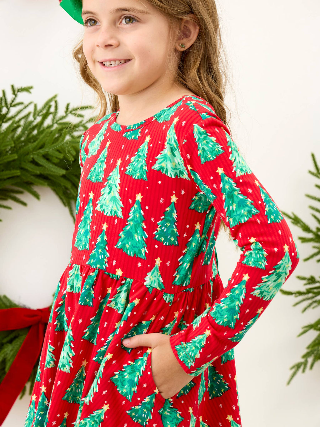 A young girl wearing a red long-sleeve dress with a green Christmas tree pattern, standing in front of a white background with holiday greenery.