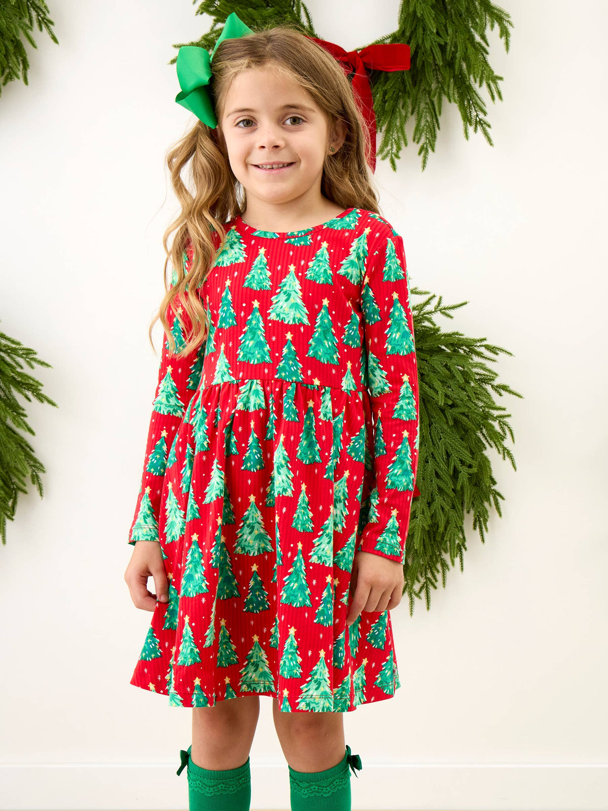 A young girl wearing a festive red long-sleeve dress with a green Christmas tree pattern, styled with a green and red bow in her hair and green socks, standing against a white wall with green pine garlands.