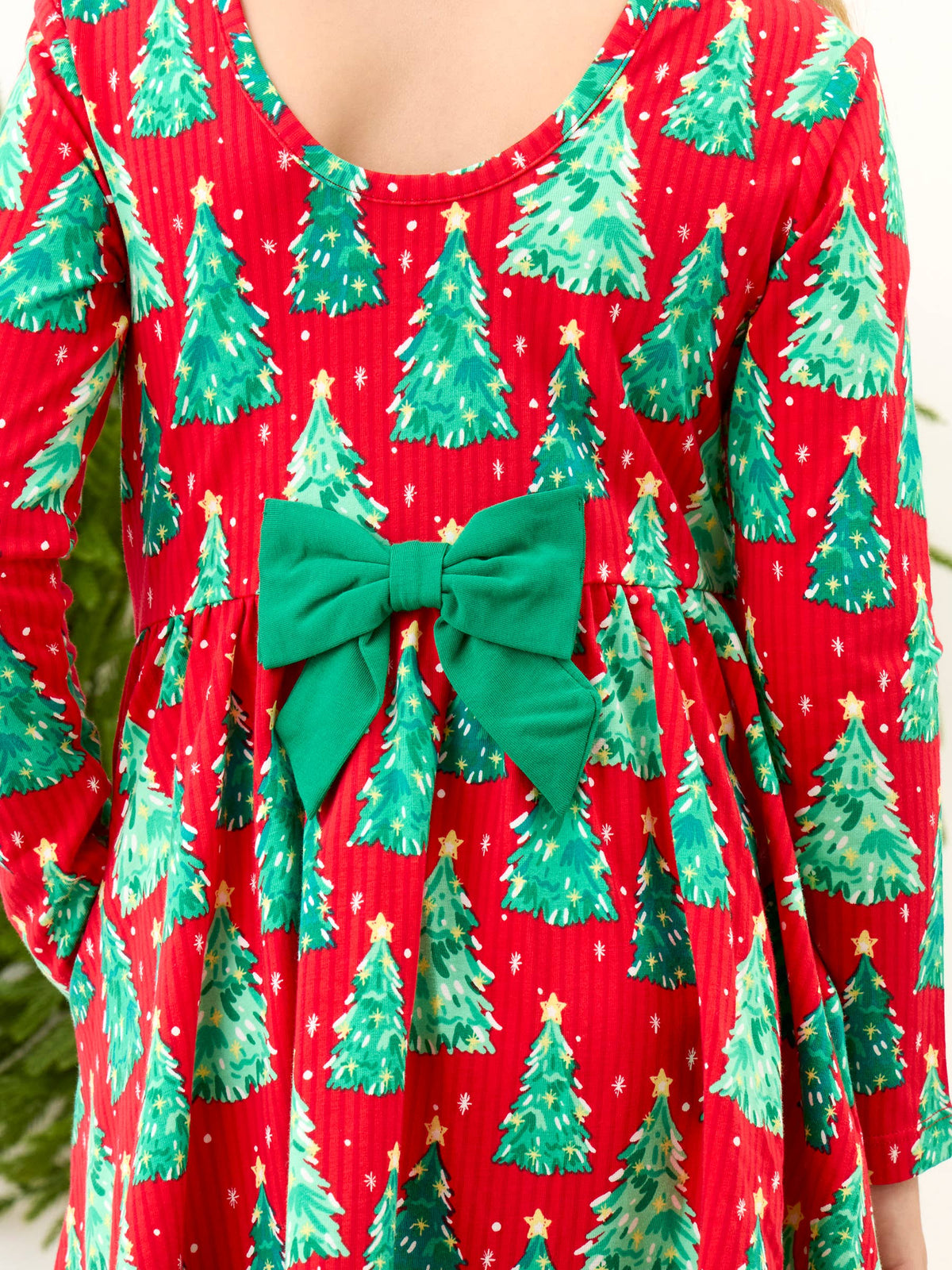 A close-up of a red long-sleeve dress with an all-over green Christmas tree print and a large green bow at the waist.