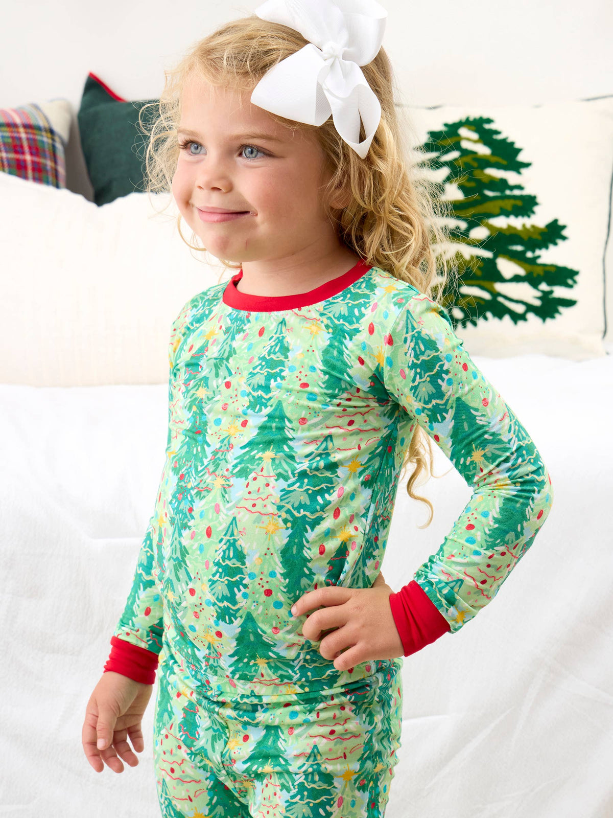 A young girl with a large white bow in her hair is wearing a long-sleeve Christmas-themed pajama shirt and matching pants. The pajamas are light green with a pattern of green Christmas trees, red trim at the collar and cuffs, and small colorful ornaments.