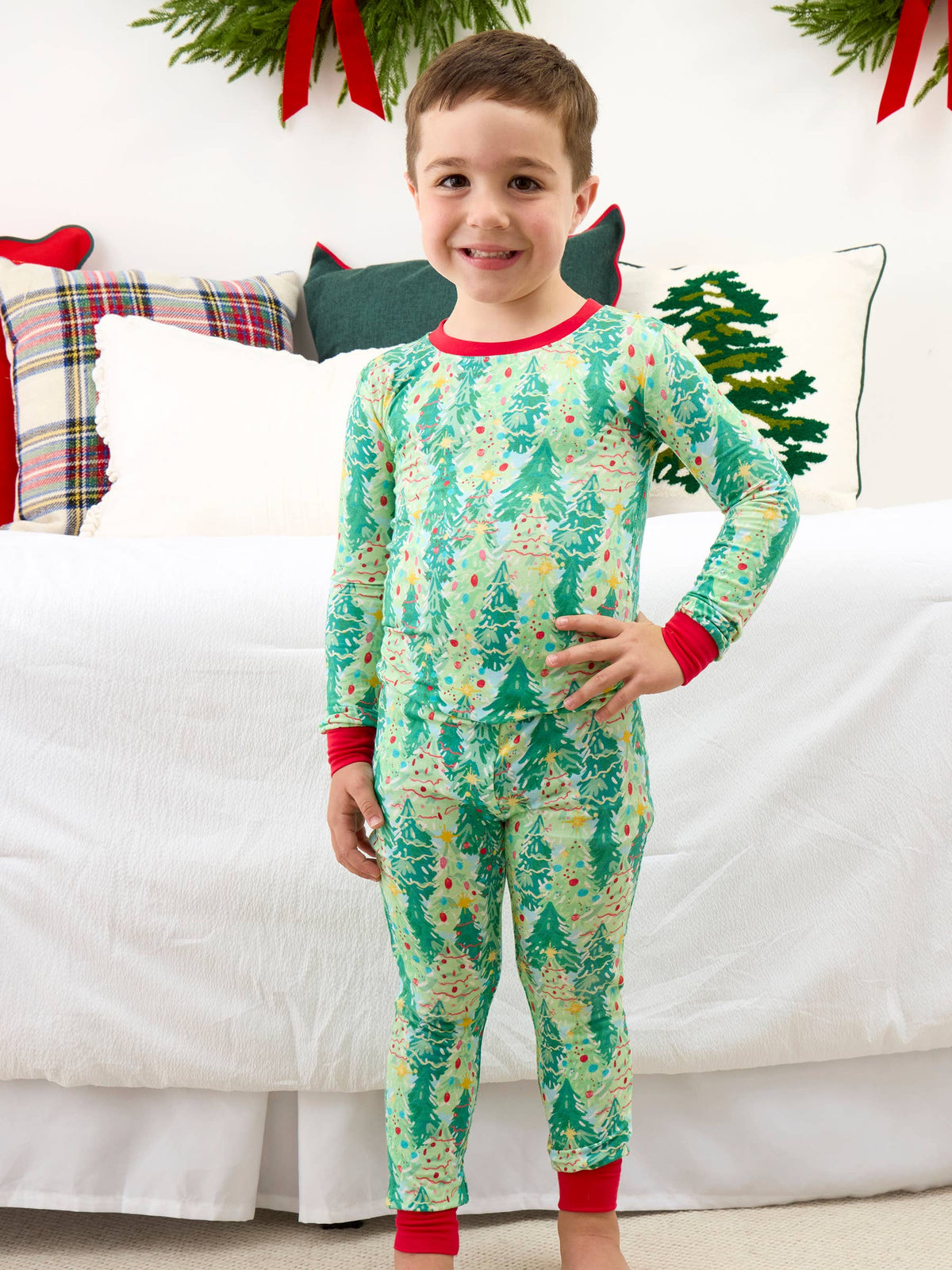 A young boy stands in front of a white bed wearing a long-sleeve Christmas pajama set with a green tree pattern and red trim.