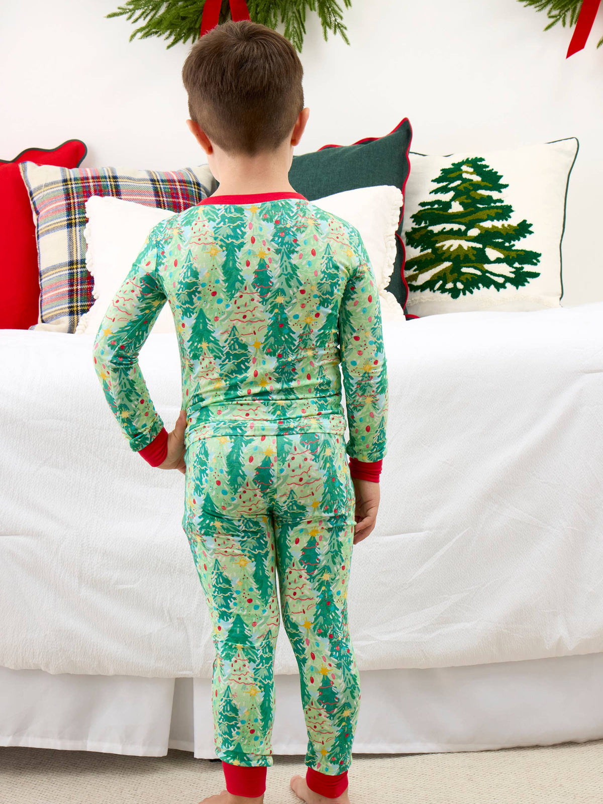 A child viewed from behind wearing a long-sleeve and matching pajama set with a green Christmas tree pattern and red cuffs and collar, standing in front of a white bed with holiday pillows.
