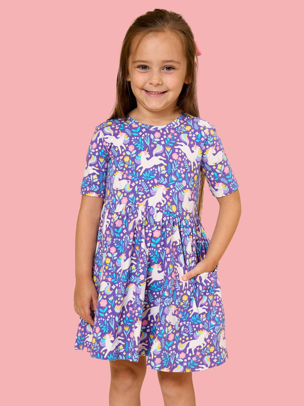 A young girl with long brown hair stands against a solid pink background wearing a short-sleeve purple dress with a white unicorn and colorful floral pattern.
