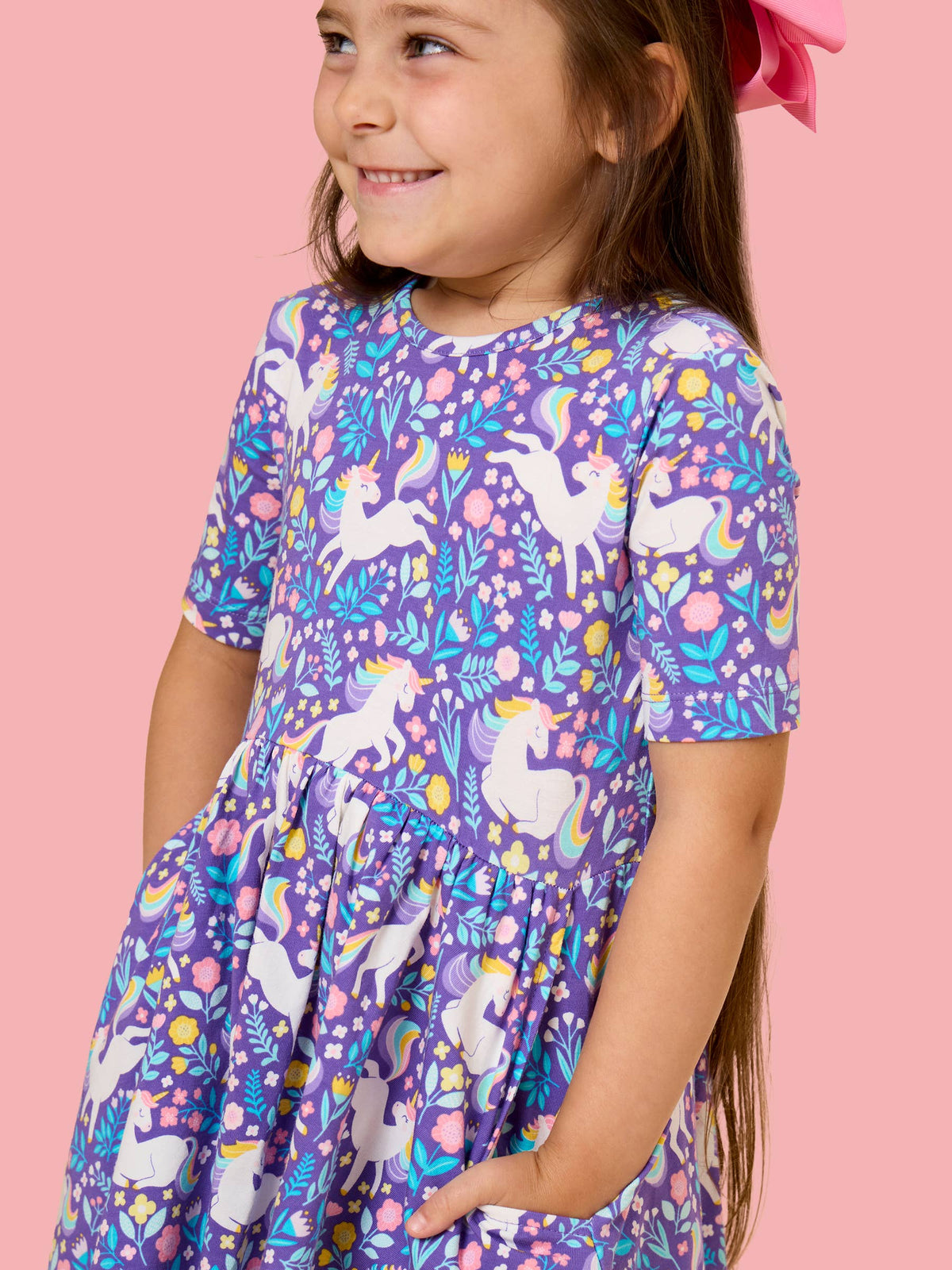 A young girl wearing a purple short-sleeve dress with a colorful unicorn and floral print, standing against a solid pink background.