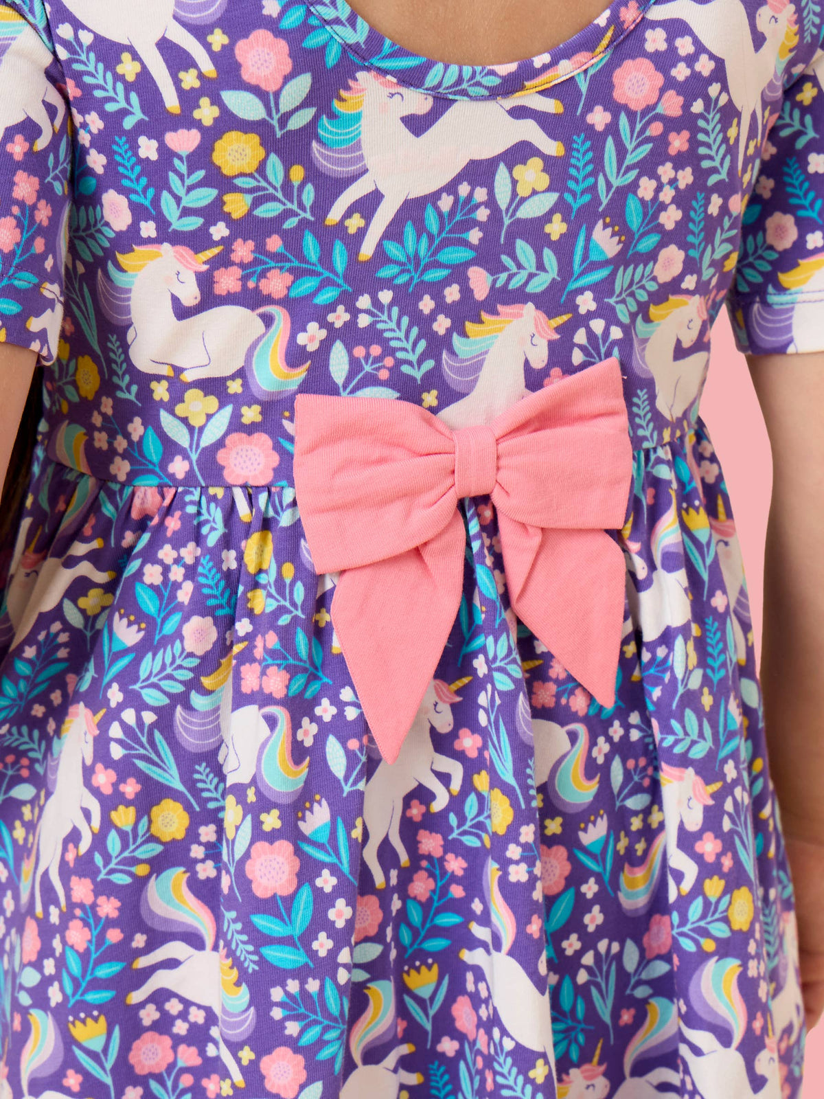 A close-up of a child wearing a purple dress with a colorful unicorn and floral print, featuring a large pink bow at the waist.