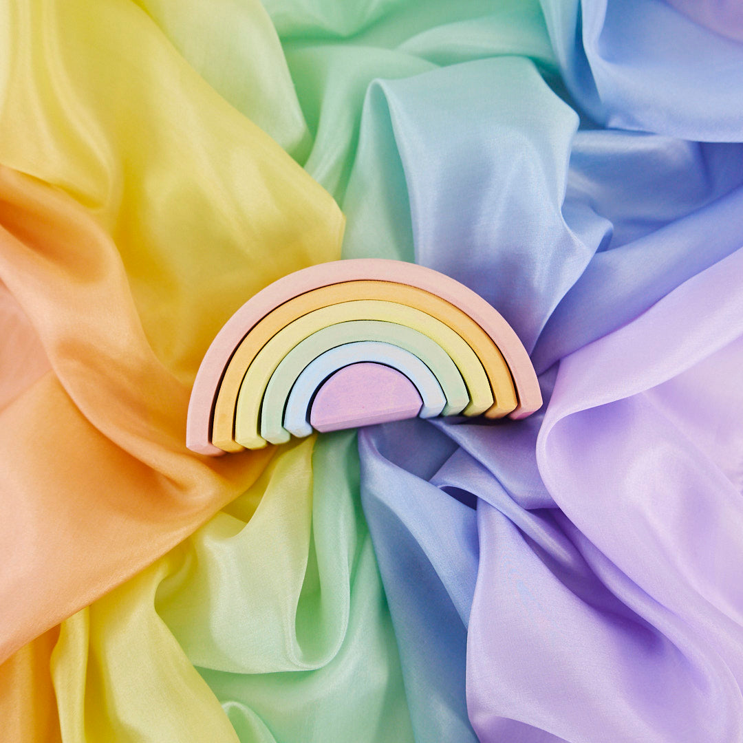 A pastel rainbow wooden toy or decorative piece with layered arches in pink, yellow, green, blue, and purple, placed on draped satin fabric in a matching rainbow color palette.