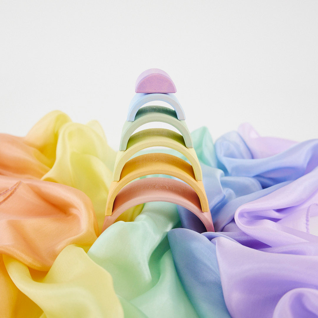 A pastel rainbow hair claw clip arranged upright in the center, surrounded by matching draped satin fabric in yellow, green, blue, and purple on a clean white background.