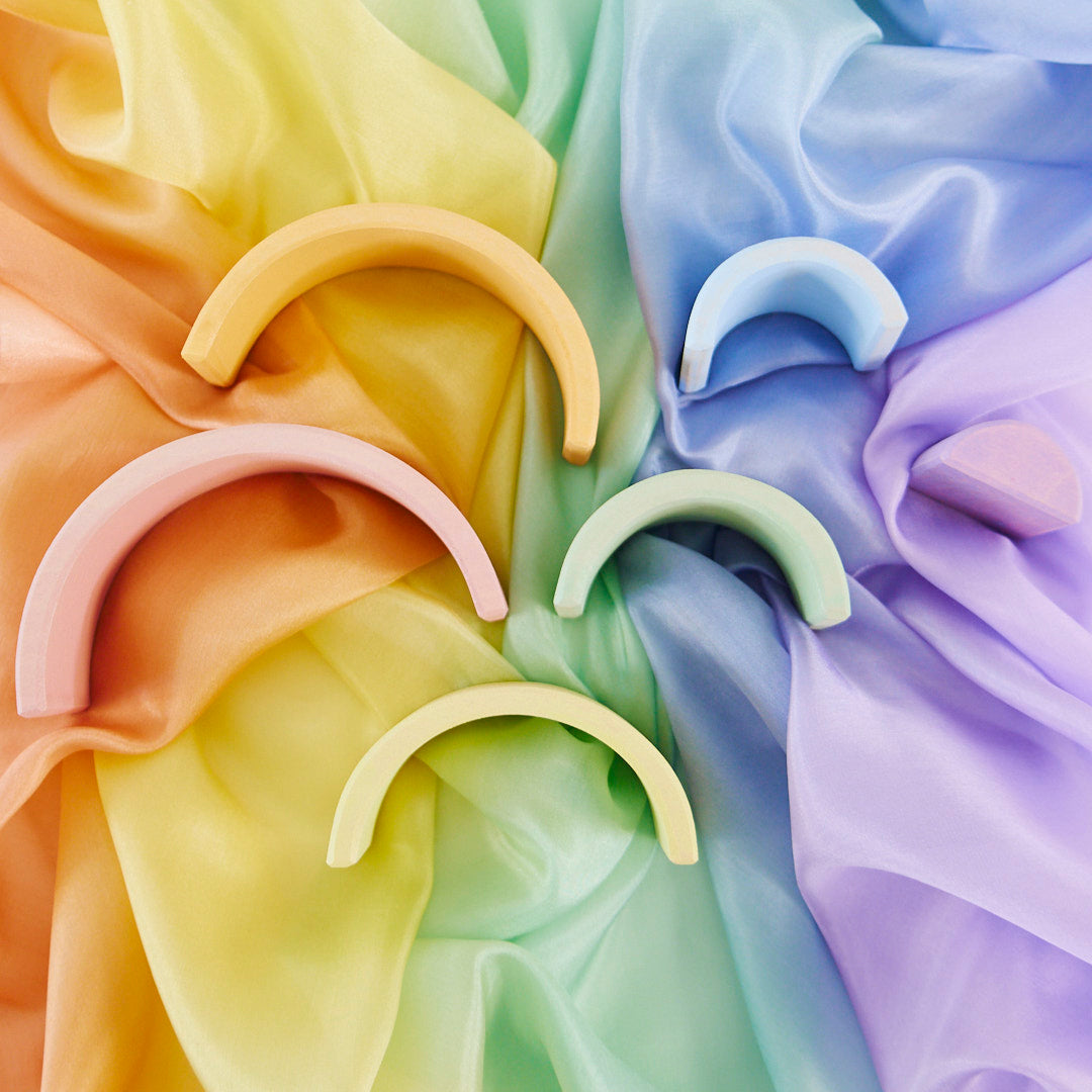 Five pastel crescent-shaped hair clips in soft peach, pink, light blue, mint green, and pale yellow, arranged on draped satin fabric with a rainbow color gradient background.