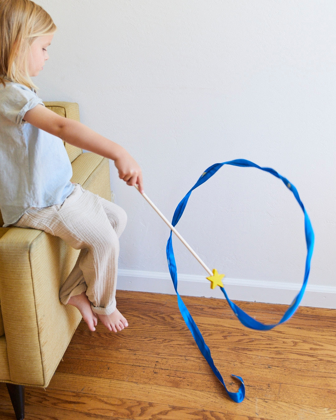 A young child sitting on a yellow armchair, holding a wooden wand with a long blue ribbon and a yellow star topper on a hardwood floor.