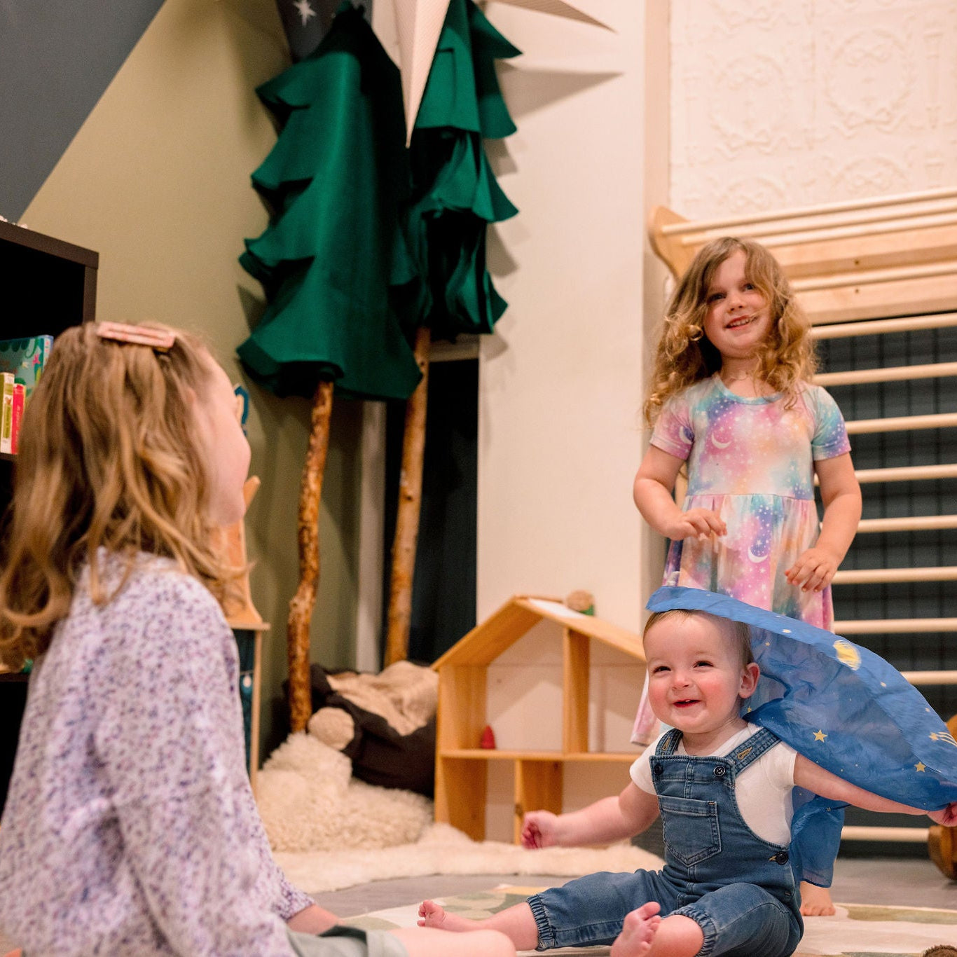 A baby in denim overalls sits smiling on a play mat in a playroom, holding a blue dinosaur toy while a toddler stands behind them in a pastel tie-dye shirt, and another child is seated in the foreground. The room contains wooden climbing bars, a bookshelf, and soft play items.