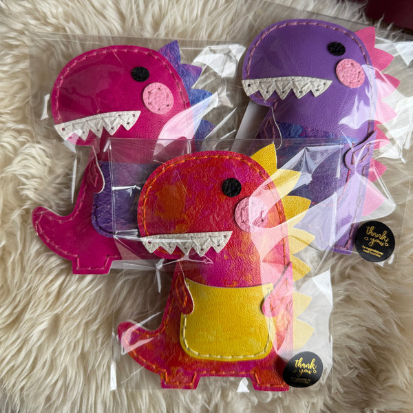 Three colorful dinosaur-shaped cushions in clear packaging on a fluffy white surface.