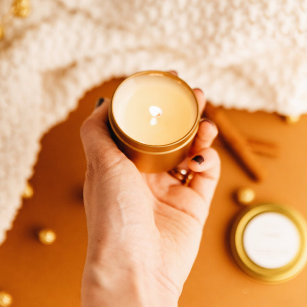 Hand holding a small gold candle with another candle in the background on a textured surface.