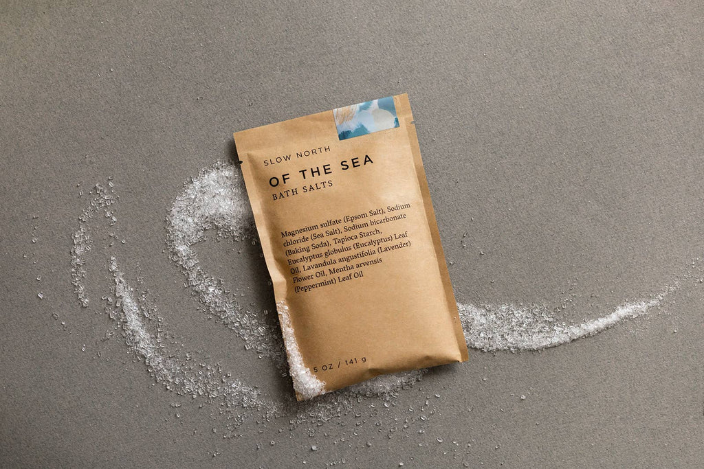 Brown package of 'Of the Sea' bath salts on a gray surface with scattered salt crystals.