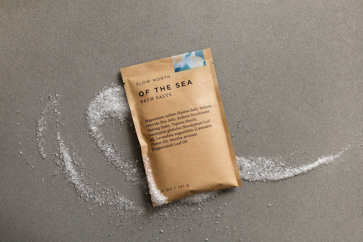 Brown package of 'Of the Sea' bath salts on a gray surface with scattered salt crystals.