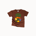 A brown short-sleeve kids' t-shirt with a colorful graphic on the front that reads 'LITTLE LOCAL' above cartoon vegetables, cheese, and a jar, shown on a light gray background.