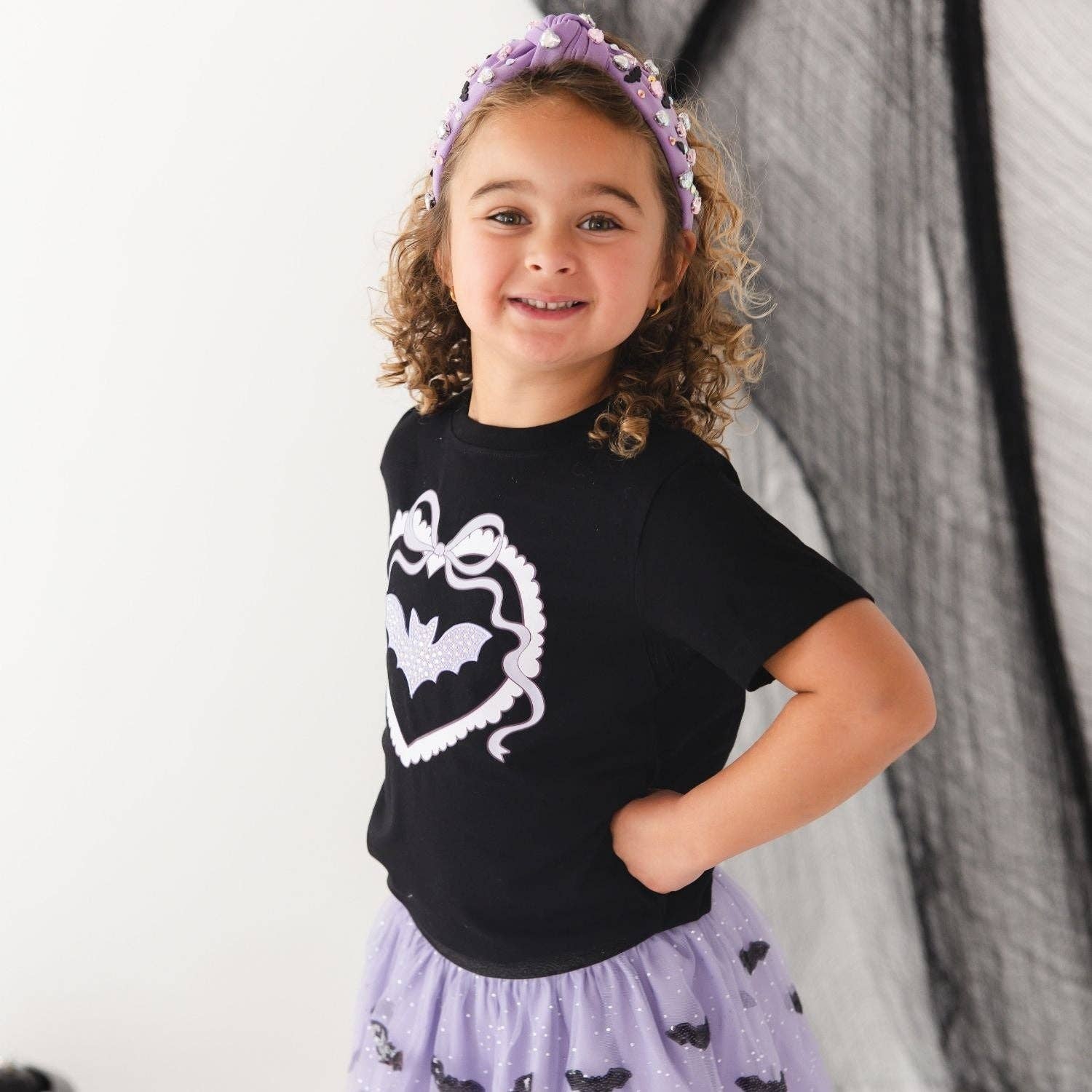 A young girl with curly brown hair wearing a black short-sleeve t-shirt with a white bat graphic, a light purple glittery skirt with bat motifs, and a matching purple glittery headband, posing against a white and black backdrop.