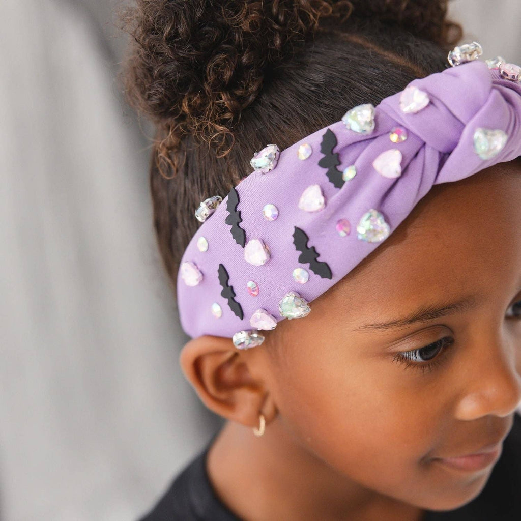 A close-up of a child wearing a lavender purple knotted fabric headband decorated with black bat silhouettes, white and clear rhinestone hearts, and small iridescent gems.