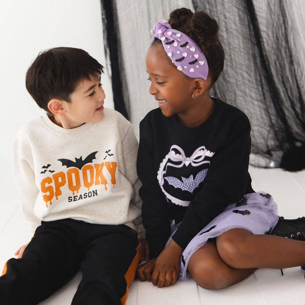 Two children sitting together in a studio setting, wearing coordinated Halloween-themed apparel. The boy wears a cream sweatshirt with orange 'SPOOKY SEASON' text and black pants, while the girl wears a black sweatshirt with a white bat graphic and a lavender bat-print skirt.