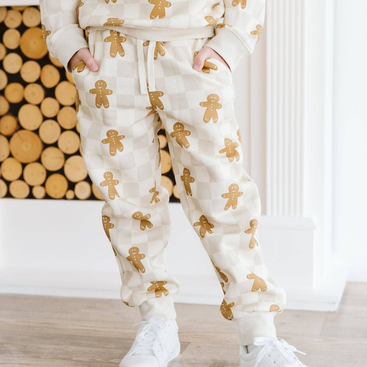 A person wearing cream-colored checkered sweatpants with a gold gingerbread man pattern, paired with a matching cream sweatshirt and white sneakers, standing in front of a wood log wall.