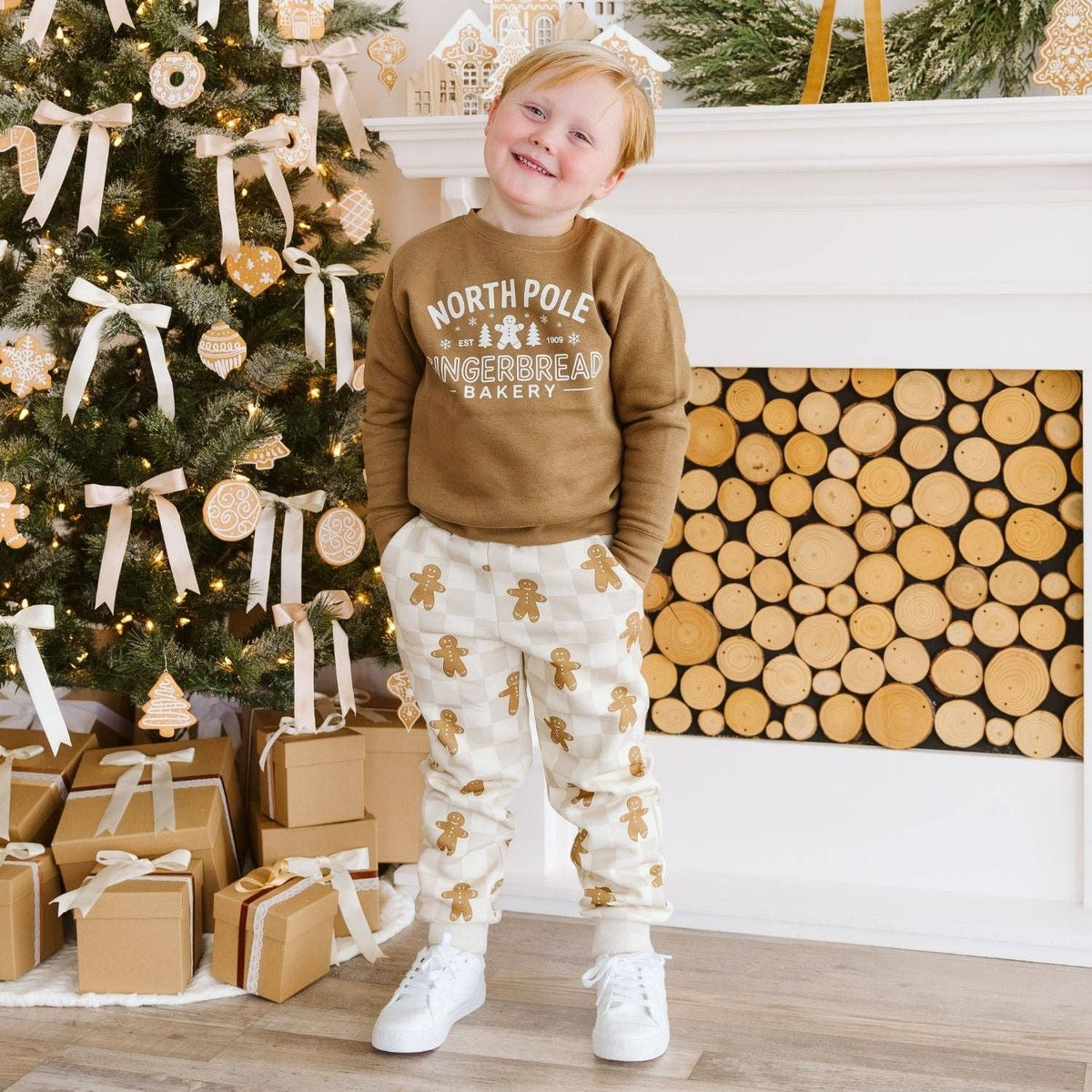 A young child wearing a brown sweatshirt with 'NORTH POLE GINGERBREAD BAKERY' printed on the front and cream pants with gingerbread man patterns, standing in front of a decorated Christmas tree and fireplace.