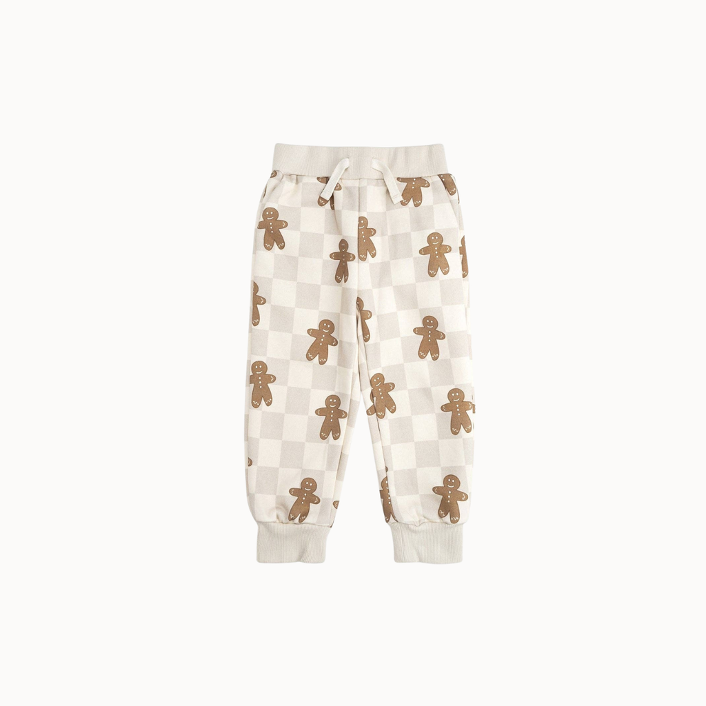 A pair of cream-colored sweatpants featuring a checkerboard pattern and an all-over print of brown gingerbread men, displayed against a solid off-white background.