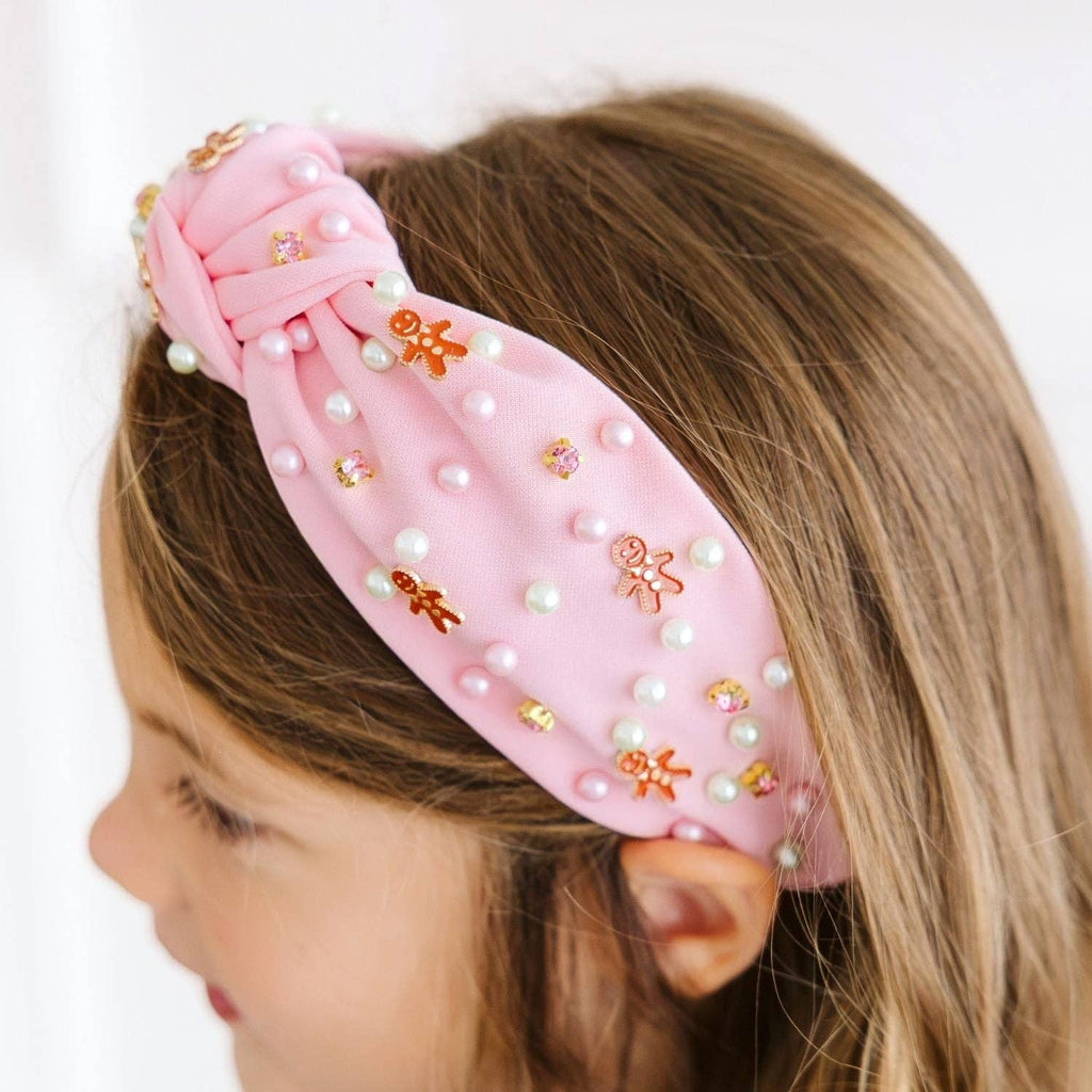 A close-up side view of a young girl wearing a light pink fabric headband decorated with white faux pearls, small gold and orange gingerbread cookies, and rhinestones.
