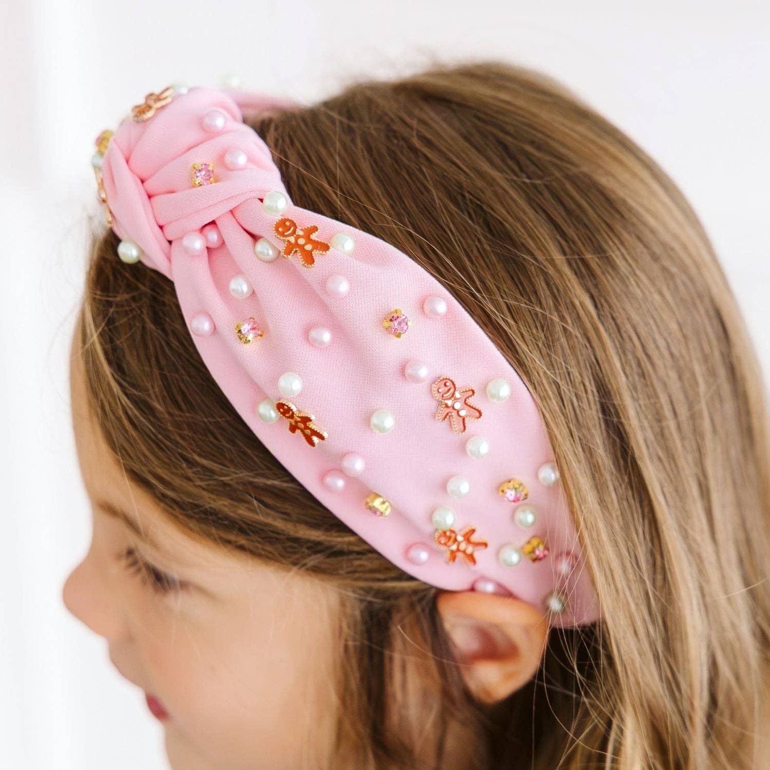 A close-up side view of a young girl wearing a light pink fabric headband decorated with white faux pearls, small gold and orange gingerbread cookies, and rhinestones.