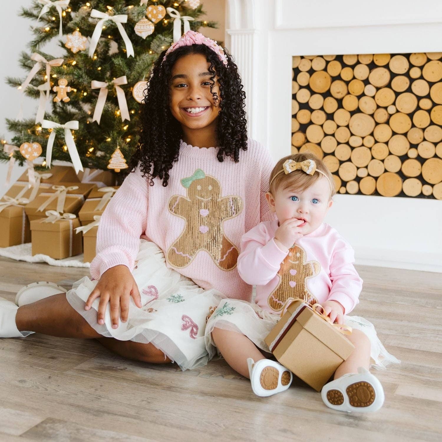 A young girl and a baby sit on a wooden floor in front of a decorated Christmas tree and a wood-burning fireplace. Both are wearing matching light pink knit sweaters with a gingerbread man graphic, white tulle holiday skirts, and matching pink and white booties.