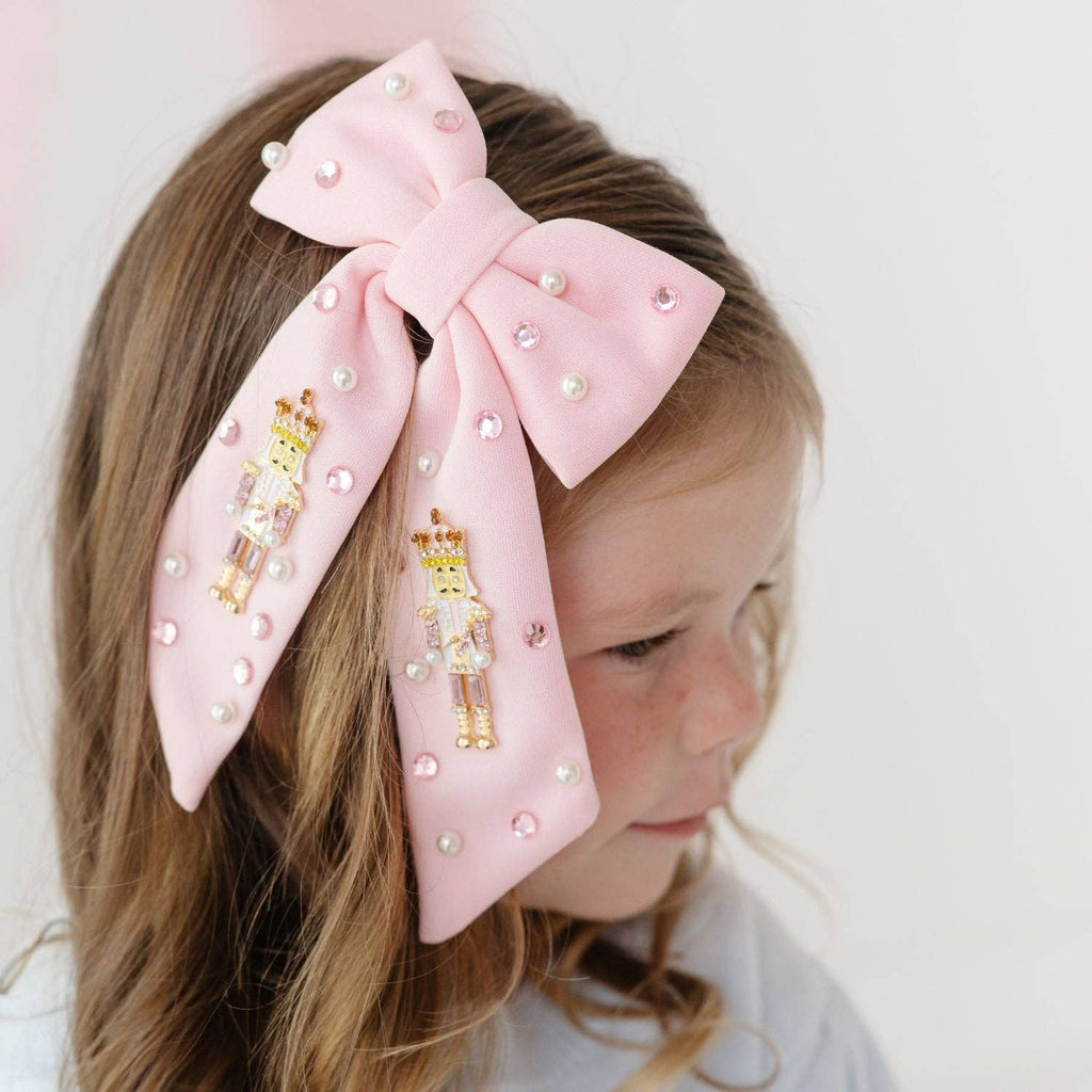 A close-up of a young girl wearing a large light pink bow hair clip adorned with small pearl-like beads, pink rhinestones, and two gold nutcracker figures, shown against a soft white background.