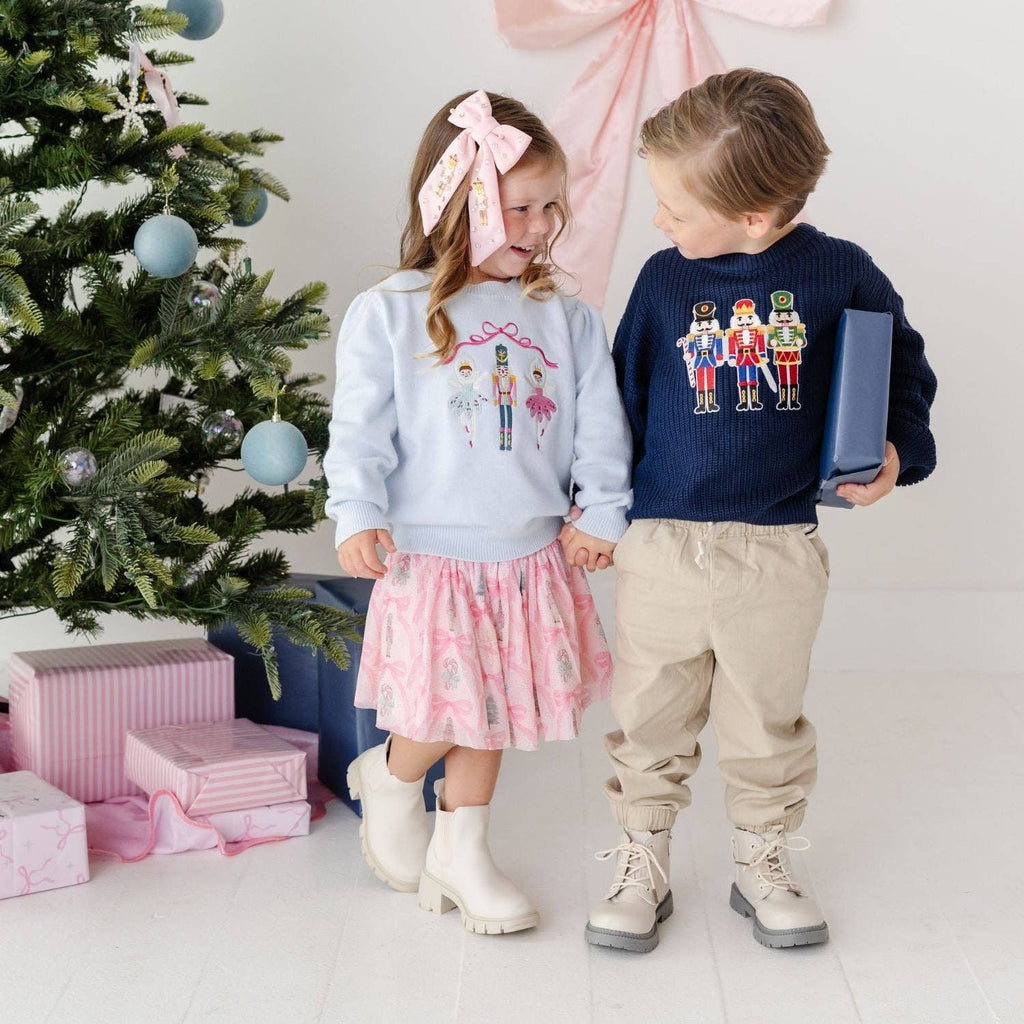 Two children standing in a holiday-themed room next to a decorated Christmas tree, wearing matching sweaters. The girl wears a light blue sweater with a pink bow headband and a pink patterned skirt, while the boy wears a navy blue sweater with nutcracker graphics, beige pants, and holds a blue gift box.