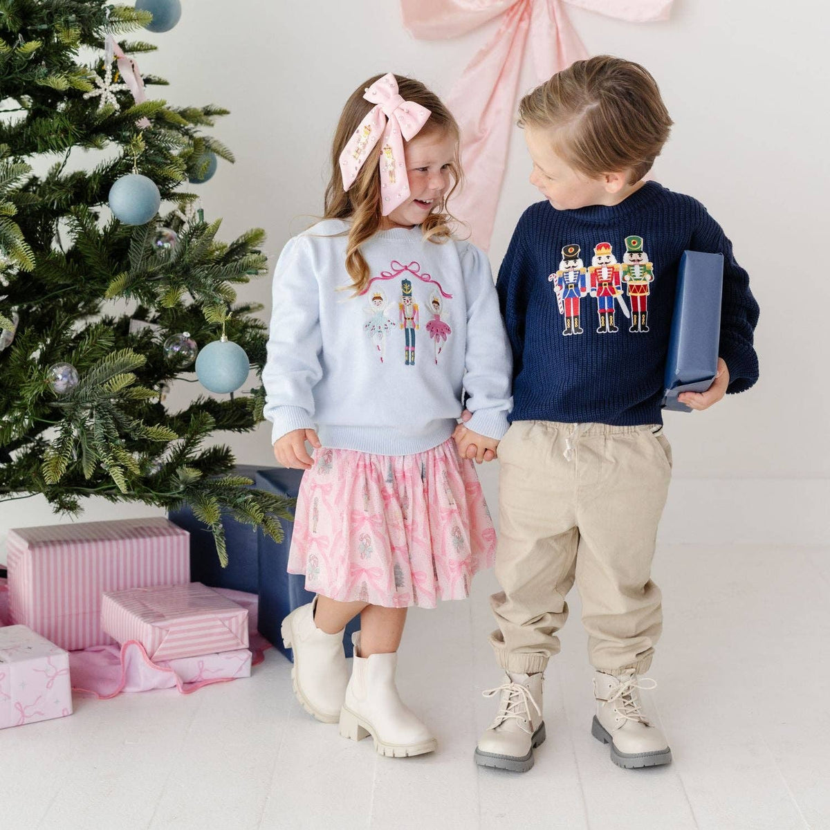 Two children standing in a holiday-themed room next to a decorated Christmas tree, wearing matching sweaters. The girl wears a light blue sweater with a pink bow headband and a pink patterned skirt, while the boy wears a navy blue sweater with nutcracker graphics, beige pants, and holds a blue gift box.