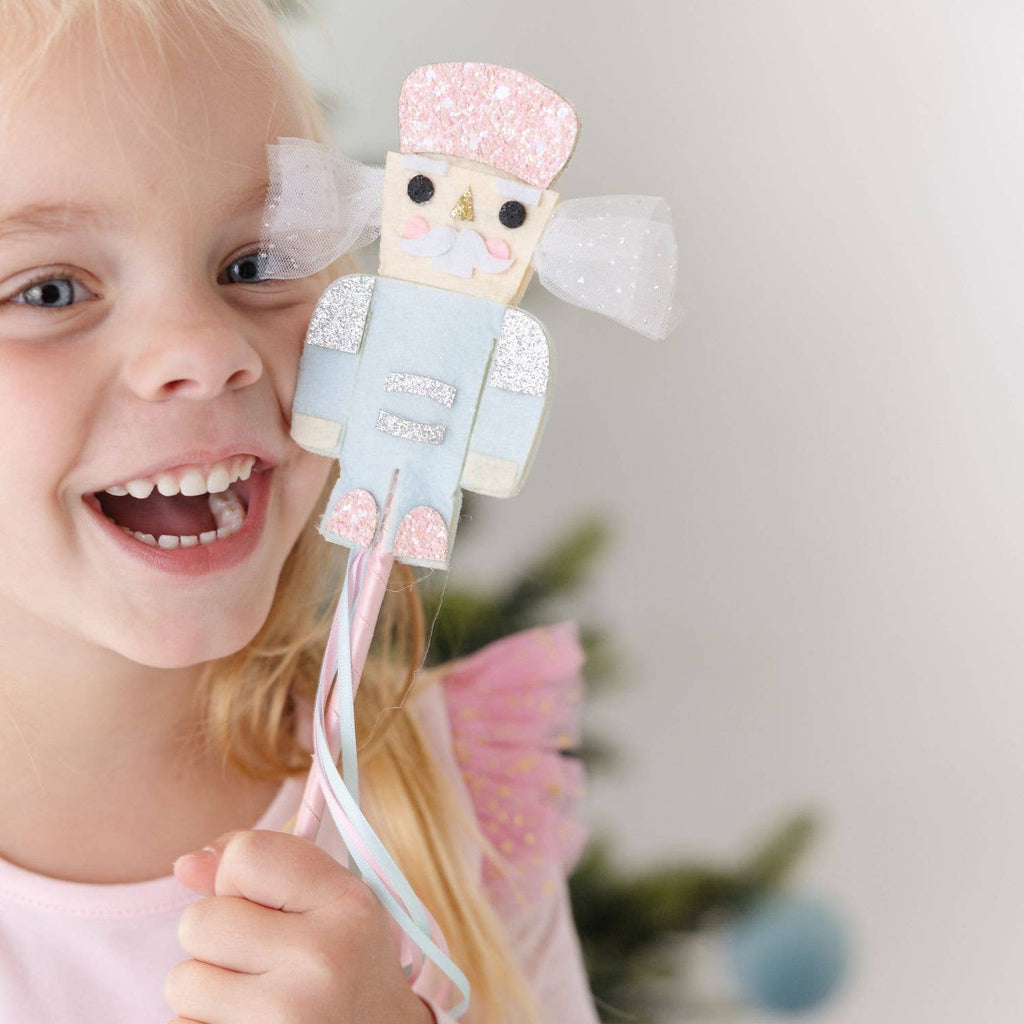 A young girl smiling and holding a nutcracker-themed finger puppet with glittery pink and light blue fabric, white tulle wings, and pastel ribbons on a wand, against a soft blurred holiday background.