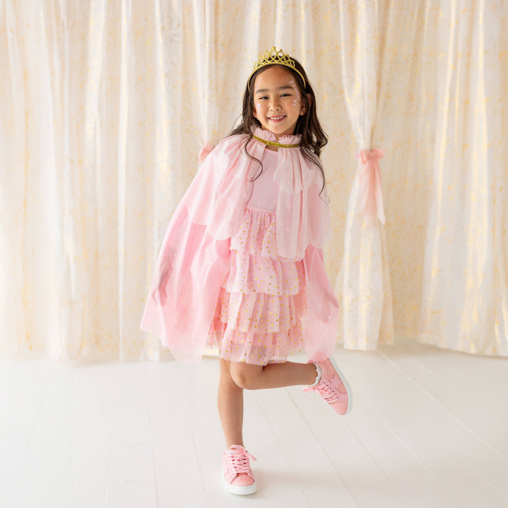 Young girl in a pink dress and tiara standing in front of a white curtain.