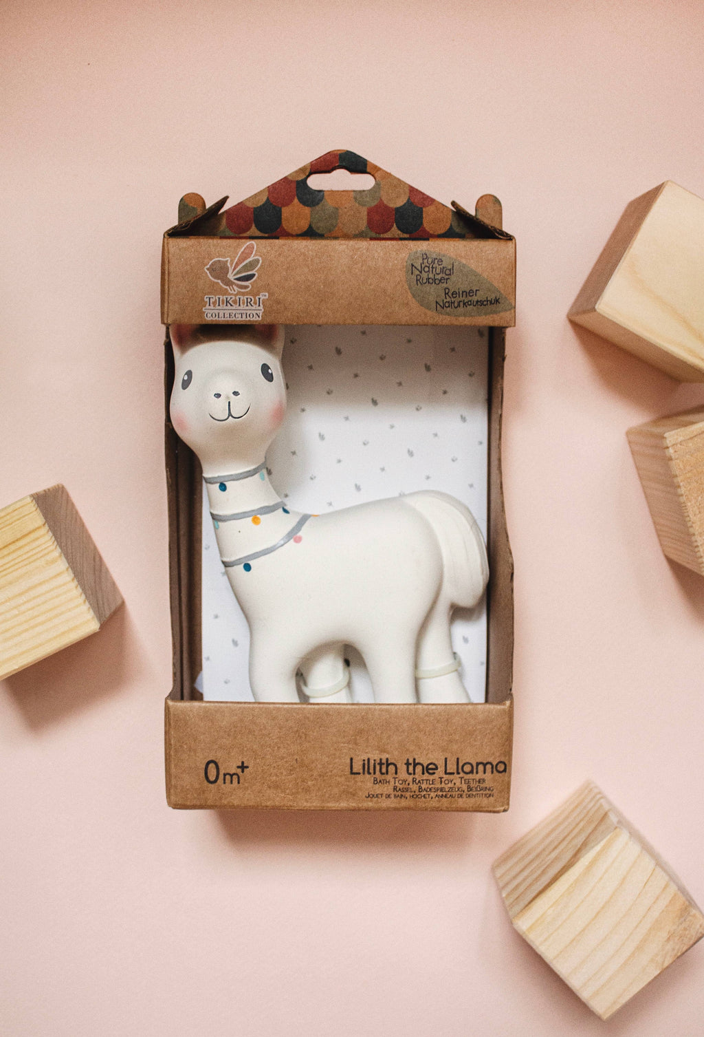 A white ceramic llama toy inside its brown cardboard packaging, displayed on a soft pink background with decorative wooden blocks.
