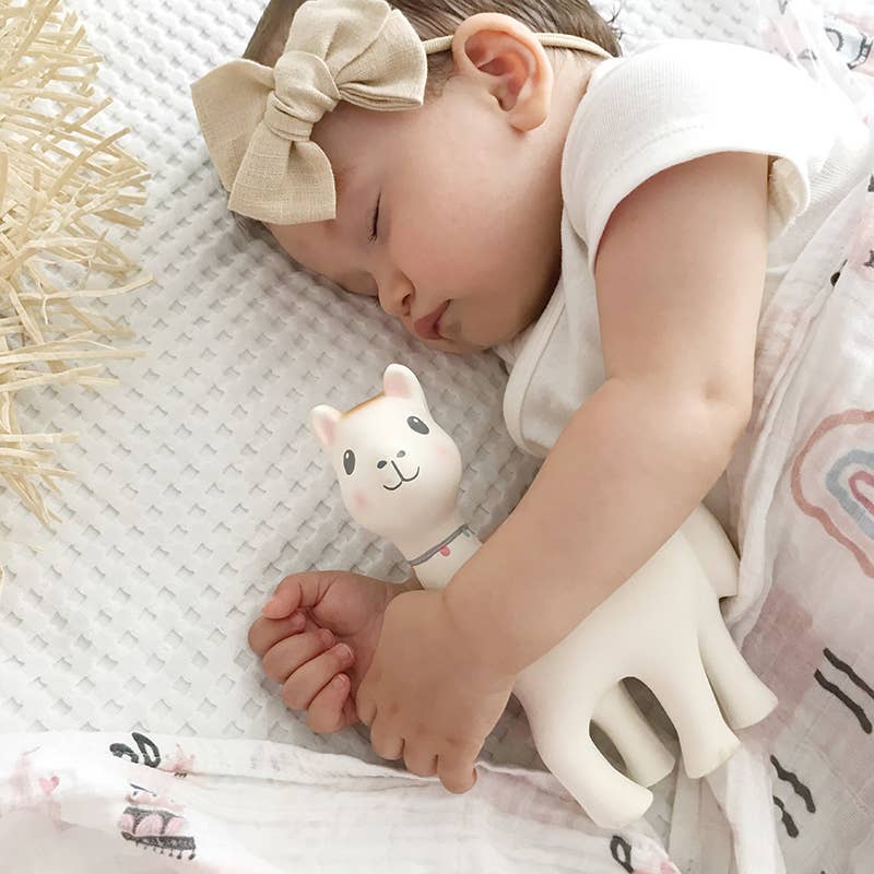 A sleeping baby wearing a beige bow headband and white sleeveless top, holding a small white llama-shaped teething toy on a textured white blanket.
