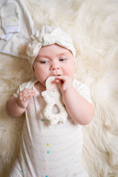 Baby wearing a white outfit with colorful stars, lying on a fluffy white surface and holding a teething toy.