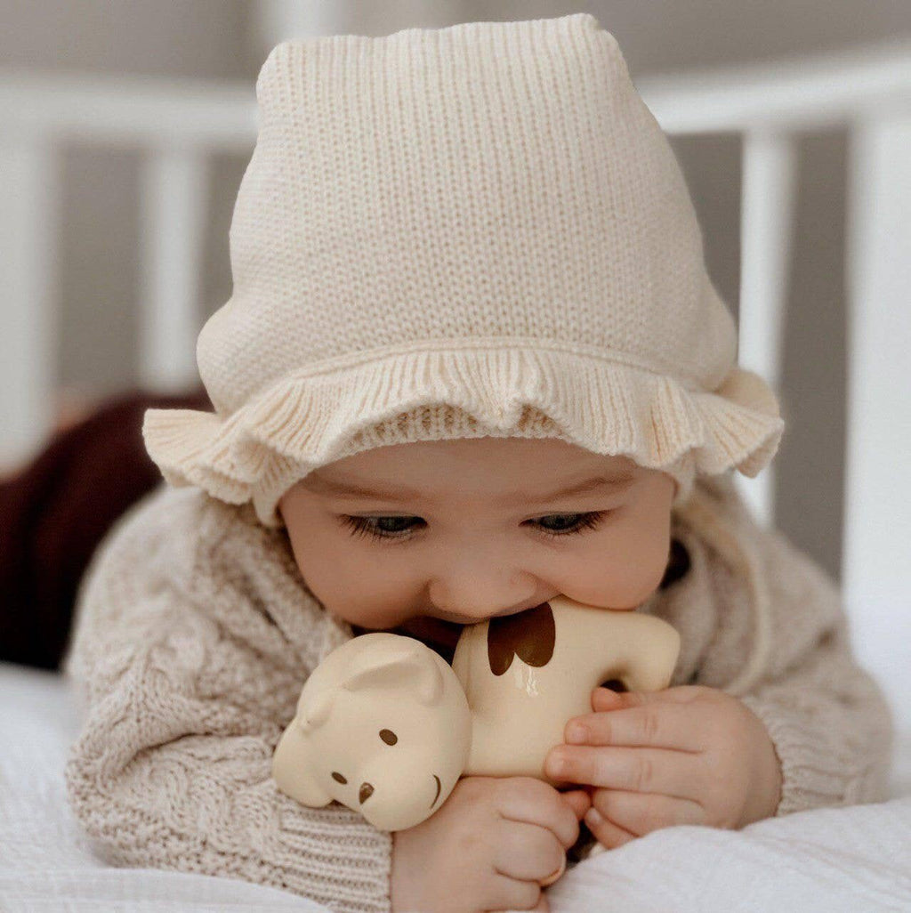 Baby wearing a beige knit hat and sweater, holding two beige animal-shaped toys.