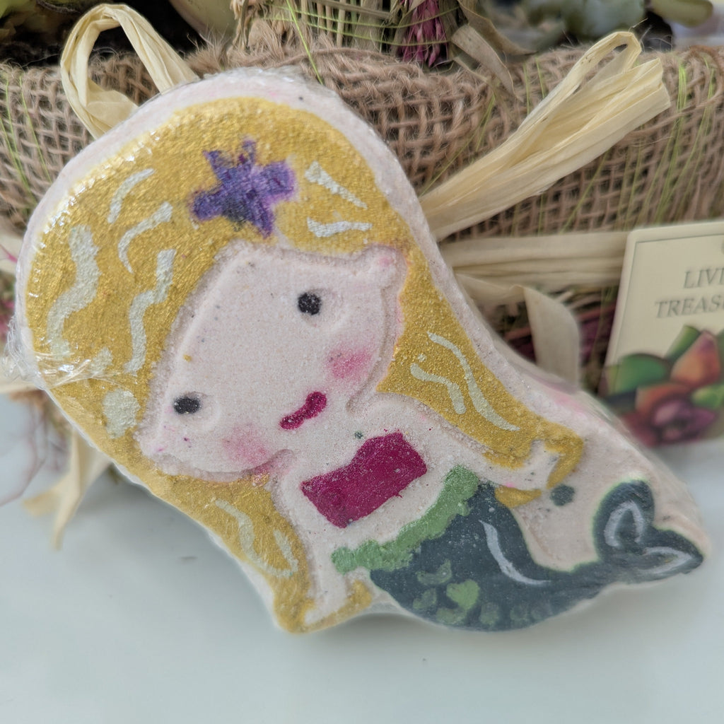 Mermaid-shaped bath bomb with blonde hair and a purple bow on a white surface.