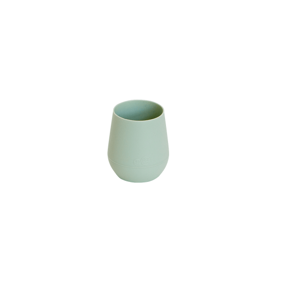 A young child wearing a navy dress and a white hairband is drinking from a green EZPZ silicone cup.